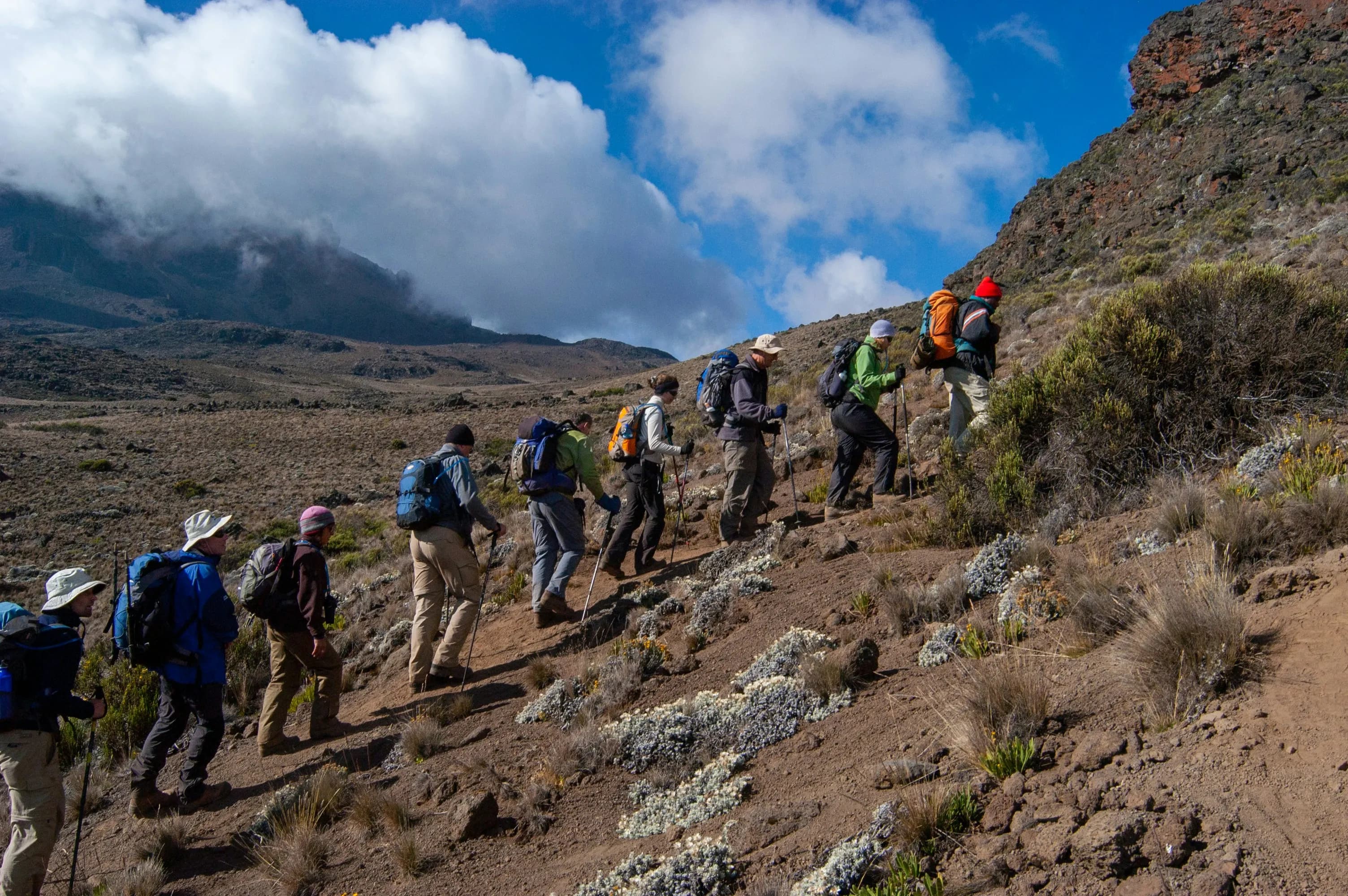 A line of trekkers with packs climbing a rocky mountain ridge below cloud.
